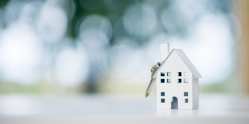 A miniature model house is sitting on a desk. A house key is dangling from the roof. The scene symbolizes purchasing a new house.